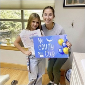 Two girls smile and pose with a sign reading No Cavity Club, promoting dental health and cavity prevention.